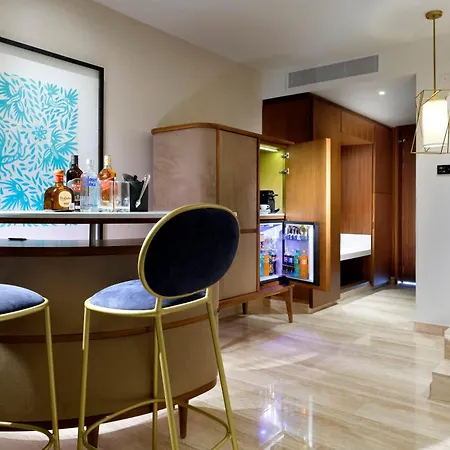 Trs Coral Hotel (Adults Only) Cancun Interior photo The photo shows a modern and stylish hotel room or suite with a well-appointed bar area. The bar is a sleek, light-colored counter with a dark wood base, set against a light beige wall. Two high-backed bar stools with dark blue velvet seats and gold 
